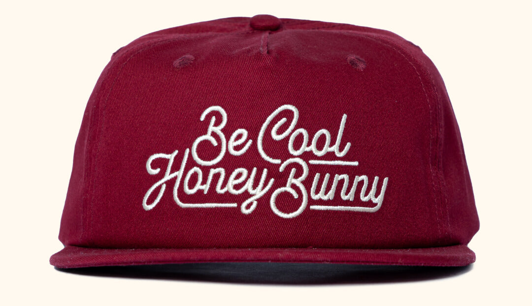 Burgundy snapback cap with cream text 'Be Cool Honey Bunny' embroidered on the front