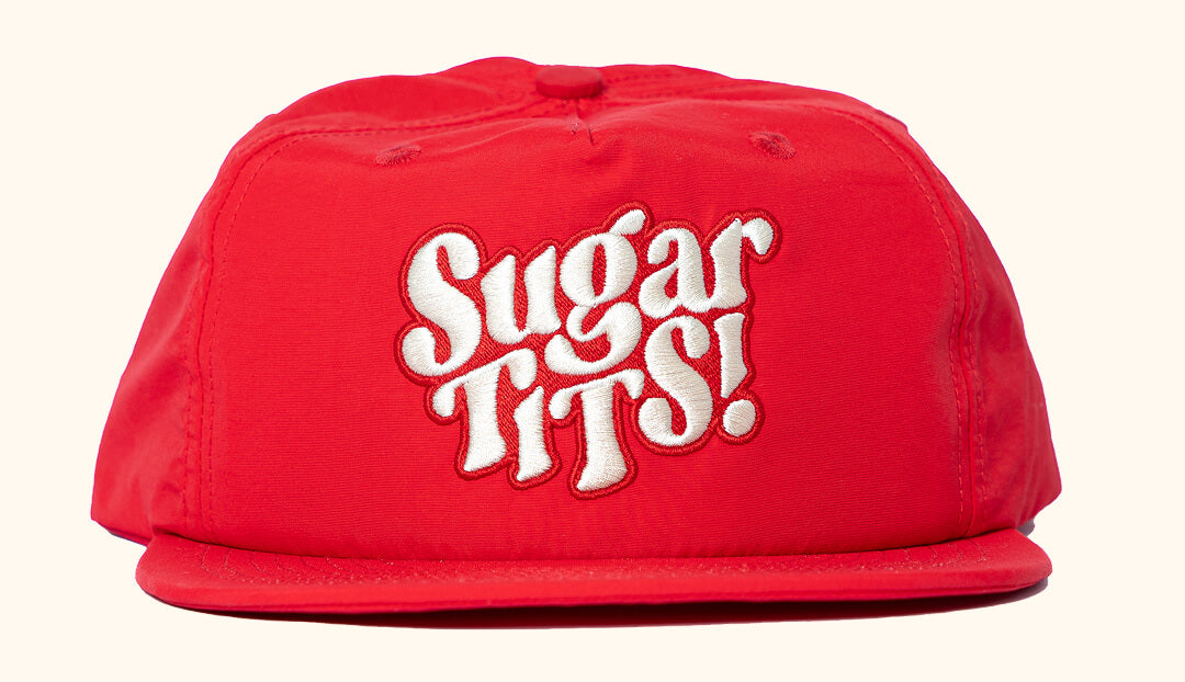 Red snapback cap with cream text 'Sugar Tits' embroidered on the front