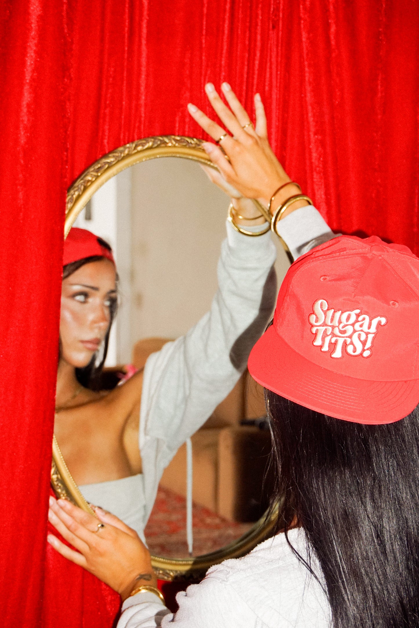 Female wearing a red cap with 'Sugar Tits!' text, looking into a mirror against a red curtain


